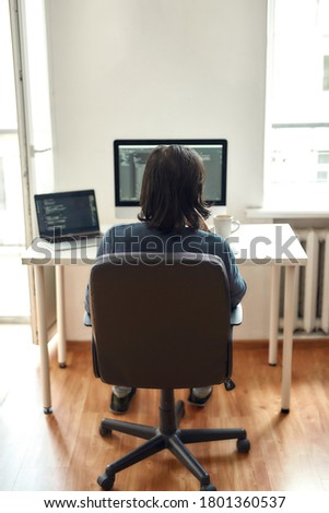 Back view of male web developer writing code while sitting at his workplace and working from home, writing code. Using desktop computer and laptop. Freelance and home office. Stay home, self isolation