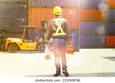 Back View Male Worker In A Helmet Holding A Toolbox And A Gallon Of Oil Walks To A Parked Forklift Truck To Inspect The Diesel Forklifts At The Workshop In The Repair Department. Maintain.