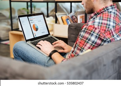 Back View Of Man Analyzes Performance On Laptop And Sitting On Sofa In Office. Side View, Coworking