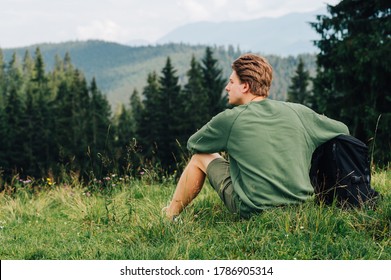 Back view a man with a backpack sits on a mountain plain among forest trees, looking away. Hipster tourist resting sitting on a meadow in the mountains, looking away