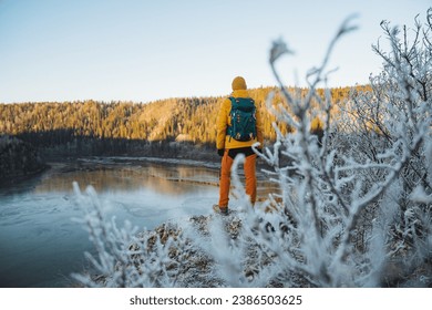 Back view of a man with a backpack standing on the edge of a cliff against the background of mountains, a guy is traveling alone, a man is standing on a mountain, looking forward at a pond