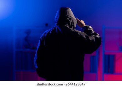 Back view of a man in  black hoodie standing in dark neon-lit room, pulling down on hoodie to cover face, evoking themes of cybersecurity, hacking, coding, and digital privacy in a tech-driven world.