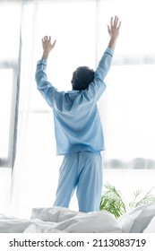 Back View Of Man In Blue Pajamas Stretching With Raised Hands Near Window