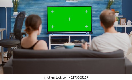 Back View Of Man Browsing Tv Channels On Green Screen Looking For Girlfriend Favourite Show While Sitting On Couch. Couple Enjoying Television Shows On Chroma Key Mockup Screen In Modern Living Room.