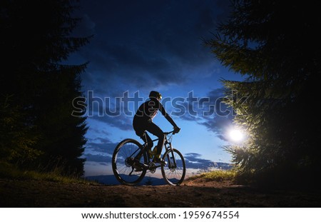 Back view of man cyclist riding bike under blue evening sky with clouds. Silhouette of bicyclist riding bicycle on the trail in night mountain forest. Concept of sport, biking and active leisure.