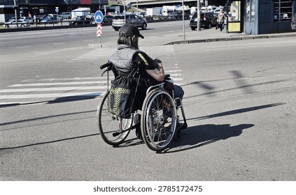 Back view of man with disability trying to cross road. Young Caucasian man in wheelchair walking in the city of Ukraine. 