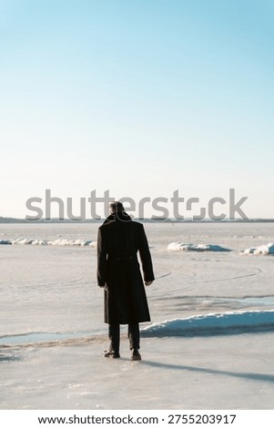 Back view of man in elegant black wool coat standing on frozen ice lake under clear blue sky in winter