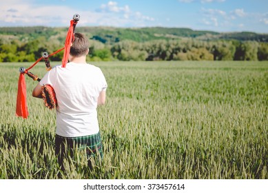 Back View Of Man Enjoying Playing Pipes In Scotish Traditional Kilt On Green Outdoors Summer Field Background