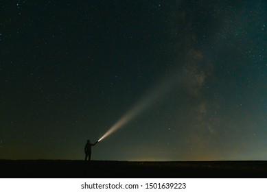 Back view of man with flashlight standing on green grassy field under beautiful dark blue summer starry sky. Night photography, beauty of nature concept. Wide panorama, copy space background.