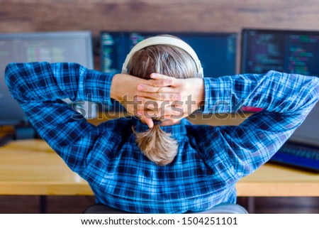 Back view of man freelancer in white headphones is working at home office. Programmer developer is writing program code software on multiple computer screens. Remote work. Geek workplace concept.