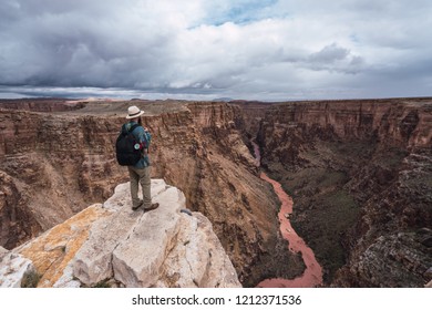 Back view of man in hat with backpack standing on peak of Grand Canyon with river in Arizona on blue sky background