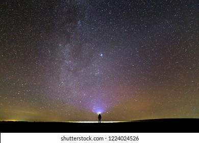 Back view of man with head flashlight standing on green grassy field under beautiful dark blue summer starry sky. Night photography, beauty of nature concept. Wide panorama, copy space background.