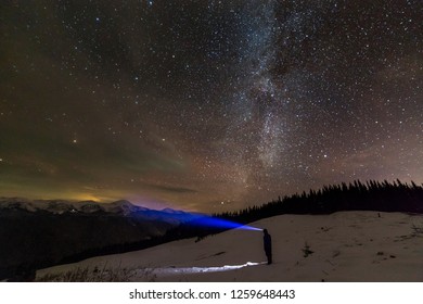 Back view of man with head flashlight standing on snowy valley under beautiful dark blue winter starry sky, bright blue beam on stars and mountains ridge background. Night photography, light painting.
