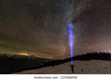 Back view of man with head flashlight standing on snowy valley under beautiful dark blue winter starry sky, bright blue beam on stars and mountains ridge background. Night photography, light painting.