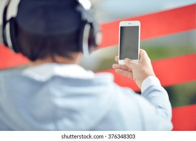 Back View Of Man With Headphones Holding Mobile Smart Phone On Outdoors Background