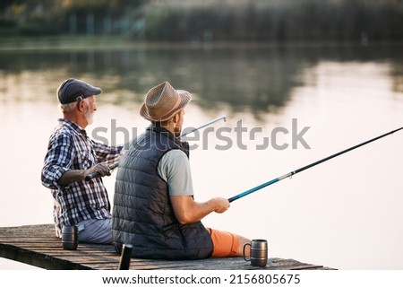Back view of man and his senior father enjoying in freshwater fishing while relaxing on a pier. Copy space.