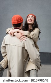 Back View Of Man Holding In Arms Joyful Woman In Beanie Hat And Trench Coat