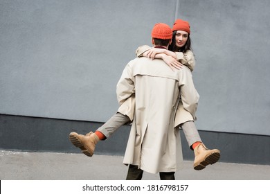 Back View Of Man Holding In Arms Woman In Beanie Hat And Trench Coat