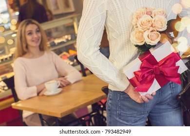 Back View Man Keeping Flower Bouquet And Gift Box Behind His Back For His Beautiful Girlfriend In Cozy Cafe. Man Prepare Unexpected Surprise For His Beloved Woman. Valentines Day, Birthday Concepts