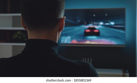 Back View, Man Playing Racing Game On Console Controlling Joystick. Young Male Gamer Plays A Car Racing Simulator Video Game.