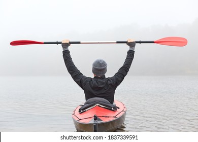 Back view of man rowing kayak across river water in foggy morning, raising his paddle up, wearing black jacket and gray cap, looks in front of him. - Powered by Shutterstock