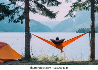 Back view of man silhouette relaxing on orange hammock between two trees pine enjoying the view at the lake in summer norwegian cloudy morning. - Powered by Shutterstock