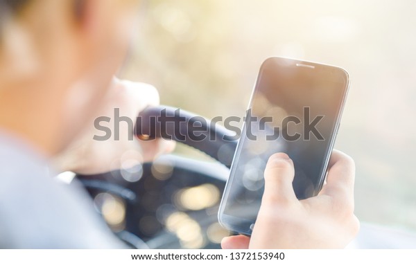 Back view of man sitting at car driver seat\
and looking at dark screen of\
smartphone
