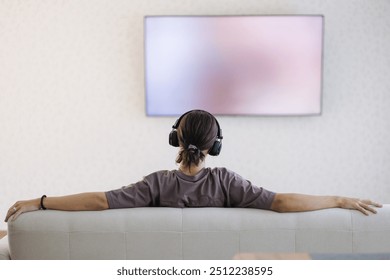 Back view of man sitting in front of big tv screen in wireless headphones on sofa - Powered by Shutterstock