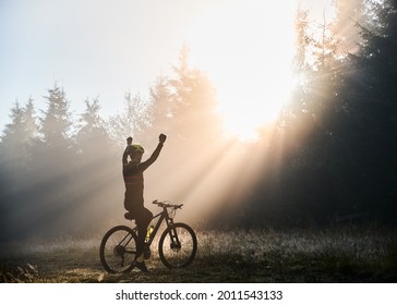 Back View Of Man Sitting On Bicycle And Raising Hands While Looking At Morning Sunlight Behind Trees. Male Bicyclist Sitting On Mountain Bike In Morning Forest. Concept Of Sport, Bicycling And Winning