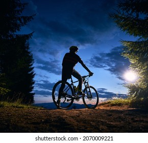 Back View Of Man Sitting On Bike Under Blue Evening Sky With Clouds. Silhouette Of Male Bicyclist Riding Bicycle On The Trail In Night Mountain Forest. Concept Of Sport, Biking And Active Leisure.