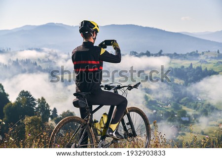 Back view of man sitting on bicycle and talking mountain photo with smartphone. Male bicyclist holding cellphone and taking picture of beautiful misty hills. Concept of sport, cycling and photography.