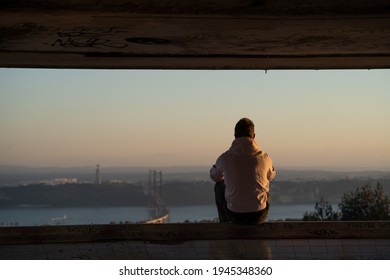 Back View Of Man Sitting And Watching Sunset Over The 25 De Abril Bridge In Lisbon. Traveler Exploring City In Wild Trip Adventure. 
