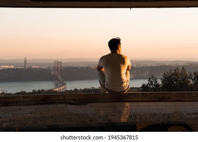 Back View Of Man Sitting And Watching Sunset Over The 25 De Abril Bridge In Lisbon. Traveler Exploring City In Wild Trip Adventure. 
