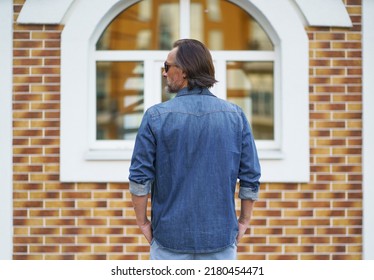 Back View Of A Man Standing Alone Looking Sideways At Old Town Building While Traveling In European Cities During Vacation Time Wearing Denim Jeans Shirt. Travel Concept. 