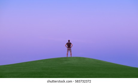 Back view of a man standing alone on the top of green hill. Evening time. Sky is blue color after sunset. Nobody around