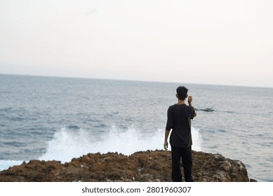 Back view of a man standing on a rocky cliff watching ocean waves crashing with white foam.