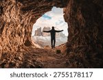 Back view of man standing with open arms at front of rocky cave Grotta delle Tre Cime with mountain peak during evening in Dolomites, Italy