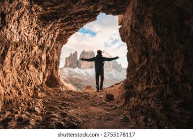 Back view of man standing with open arms at front of rocky cave Grotta delle Tre Cime with mountain peak during evening in Dolomites, Italy