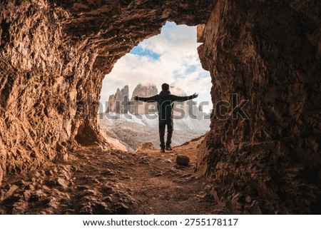 Back view of man standing with open arms at front of rocky cave Grotta delle Tre Cime with mountain peak during evening in Dolomites, Italy