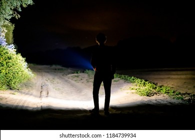 Back View Of Man Standing Outdoor On Night Forest Road Lighting The Way With Electric Flashlight