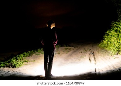 Back View Of Man Standing Outdoor On Night Forest Road Lighting The Way With Electric Flashlight