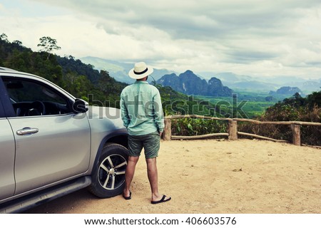 Back view of a man is thinking about this amazing world, while is standing on a mountain against jungle view. Young male traveler is enjoying beautiful landscape during road trip on suv in Thailand