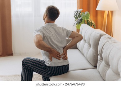 Back view of man wearing white pajamas sitting on sofa at home in the morning, touching lower back, suffering from muscle strain