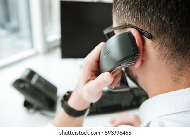 Back View Of Man In White Shirt And Eyeglasses Which Sitting By The Table With Computer And Talking On Phone. Close Up View