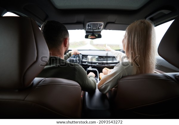 Back view of man\
and woman traveling in car. Beautiful young couple sitting on front\
passenger seats, handsome man driving comfortable automobile. Woman\
pointing on the way.