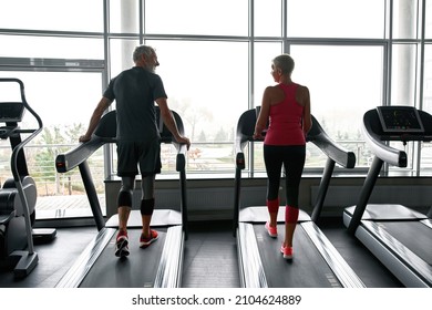 Back View Of Mature Fit Man And Beautiful Woman Run On Running Machine In Modern Gym. Workout In Fitness Center. Backlit Photo On Large Windows Background. Healthy Lifestyle And Personal Fulillment.