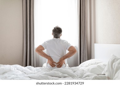 Back view of mature grey-haired man wearing white pajamas sittting on bed at home in the morning, touching lower back, suffering from muscle strain after waking up, copy space - Powered by Shutterstock