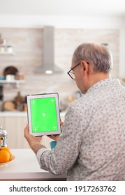 Back View Of Mature Man Holding Tablet Pc With Chroma Key In Kitchen During Breakfast. Authentic Elderly Person Using Notepad With Green Screen Isolated Mock-up Mockup For Easy Replacement