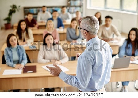 Back view of mature professor giving lecture to large group of college students in the classroom.