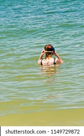 Back View, Medium Distance Of A Young Female Adjusting Mask And Snorkle Before Snorkling Near Shoreline In The Surf And Waves Of A Tropical Beach On The Gulf Of Mexico On A Sunny, Summer Day